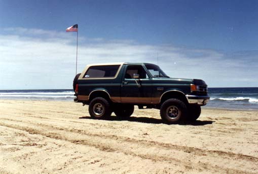 Lifted green two-tone Ford Bronco parked on a beach with an American flag and ocean behind it