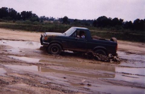 1989 Ford Bronco - A dark green Ford pickup truck navigates through a muddy, water-filled terrain, kicking up dirt and water.