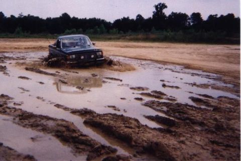 1989 Ford Bronco - A dark SUV is deeply mired in a muddy track, with trees and a dirt embankment in the background.