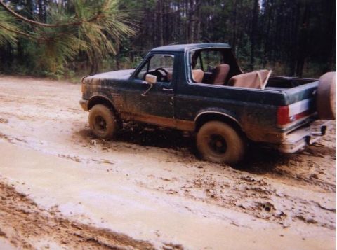 1989 Ford Bronco - A dark green Ford Bronco is shown driving through a muddy, wooded area.