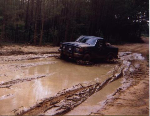 1989 Ford Bronco - A dark blue Ford Bronco navigates a muddy track surrounded by trees.