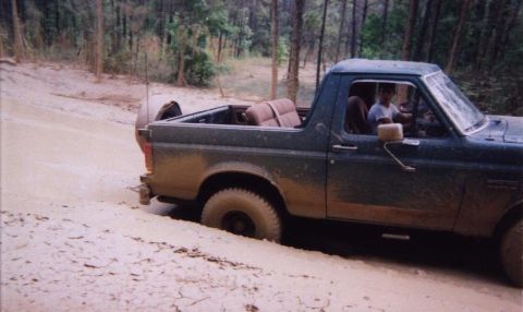 1989 Ford Bronco - A dark green Ford Bronco is stuck in a muddy, wooded area. The vehicle's tires are partially submerged in the mud.