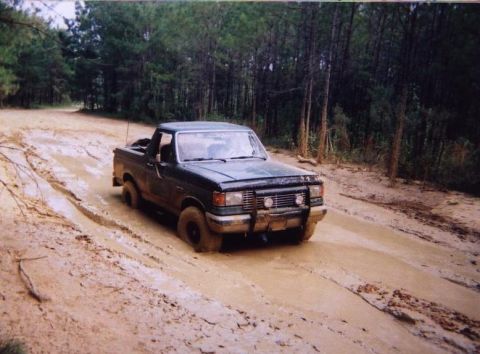 1989 Ford Bronco - A dark green Ford F-Series truck navigates a muddy trail through a forest.
