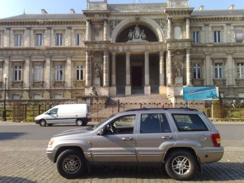 1991 Ford Crown Victoria - A silver Jeep Grand Cherokee is parked on a cobblestone street with a white van driving in the background.