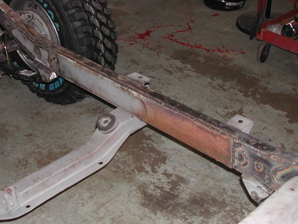 Close-up of a vehicle frame section with a tire in the background. Some rust is visible on the frame