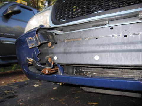 1991 Ford LTD Crown Victoria - Close-up of a damaged blue car bumper showing bent metal and rust, with another vehicle in the background.