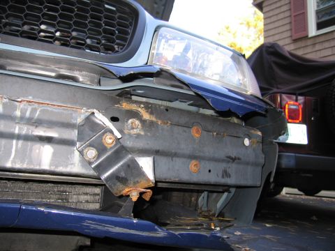 1991 Ford LTD Crown Victoria - The front end of a blue car shows significant damage to the bumper and body panels, with rust visible on the support structure.