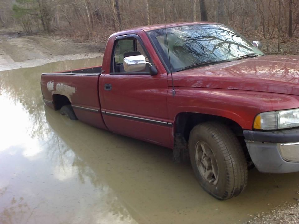 Red pickup truck with flat tire stuck in muddy water, showing rust on rear fender