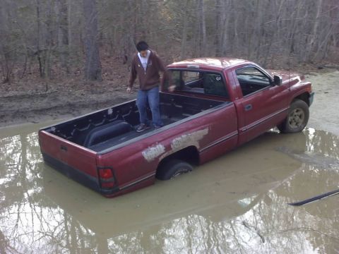1996 Dodge 1500 - A person stands in the bed of a red pickup truck that is stuck in a large mud puddle in a wooded area.