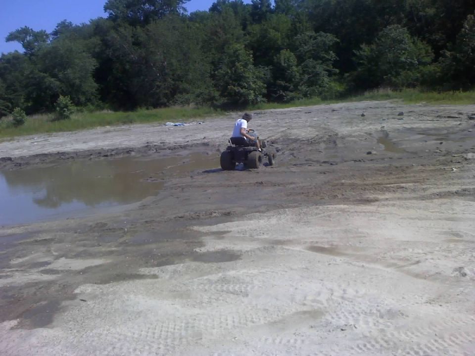 All-terrain vehicle on muddy ground near water, rider wearing white shirt