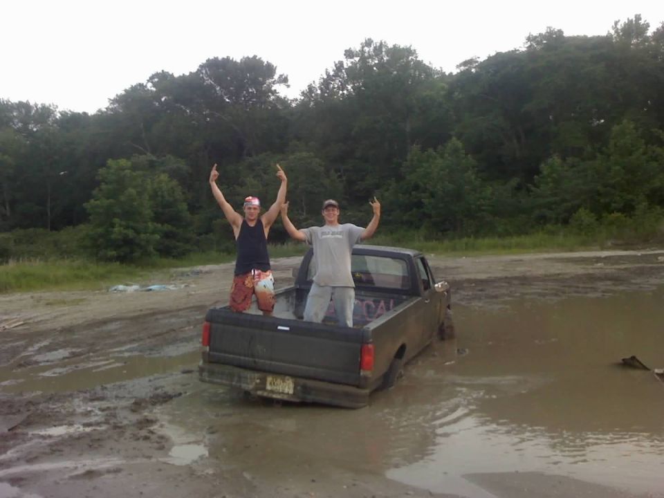 Dark gray pickup truck stuck in mud, rear view. Two men stand in the truck bed with arms raised