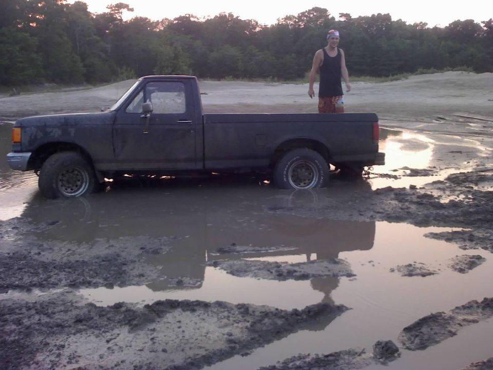 Dark pickup truck stuck in mud, side view. A man stands in the truck bed. The truck appears to be from the early to mid-1990s