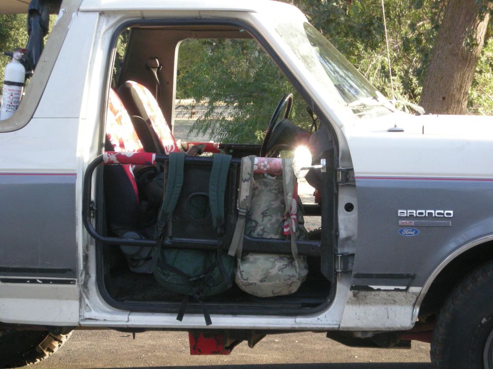 White and grey 1991 Ford Bronco side view with open door, roll cage, and backpacks inside