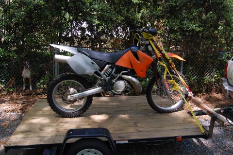 1994 Ford Bronco - A dirt bike is secured with straps to a wooden trailer, with a dog visible behind a fence.