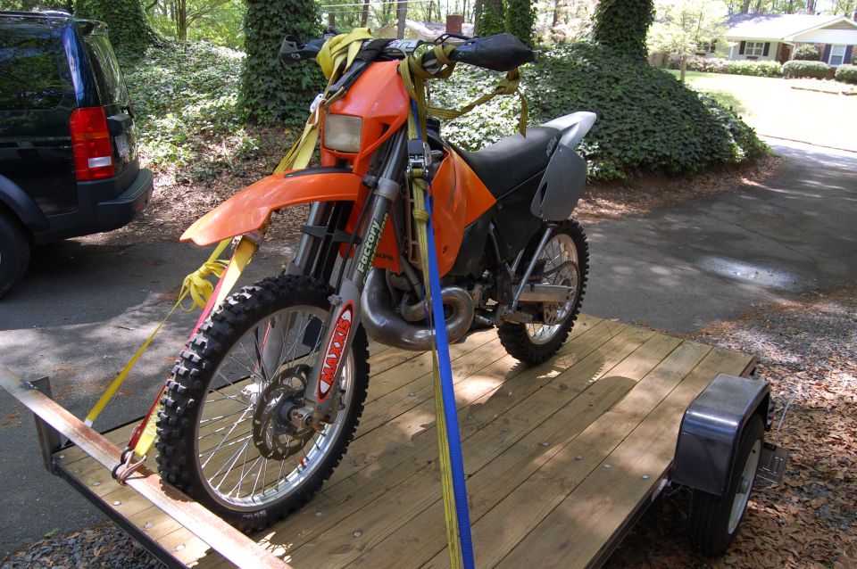 Orange dirt bike secured with yellow and blue straps on a wooden trailer. A black SUV is partially visible to the left