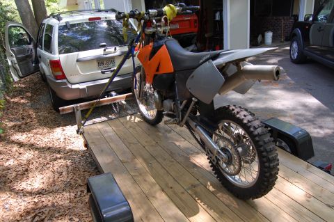 1994 Ford Bronco - A dirt bike is loaded onto a wooden trailer, towed by a white SUV with its driver's door open.