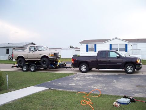 1996 Ford Bronco - A Ford Bronco is loaded onto a trailer and towed by a GMC Sierra pickup truck in a residential area.