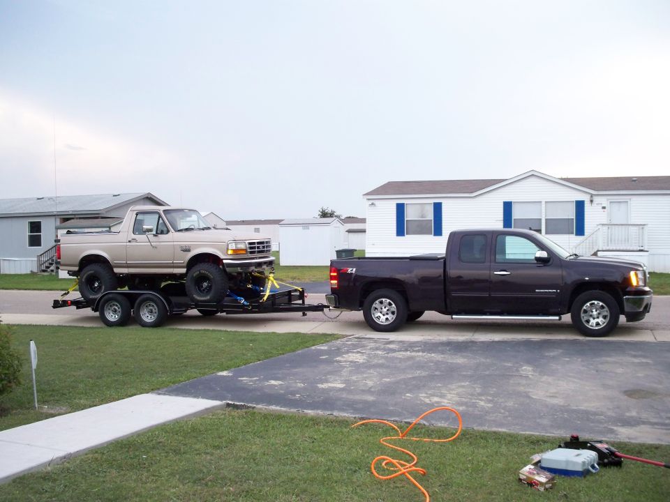 Tan Ford Bronco on a trailer, towed by a dark red GMC Sierra pickup truck