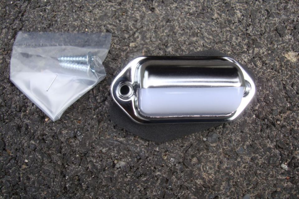 Chrome license plate light with white lens, next to a plastic bag with two screws and a small metal pin