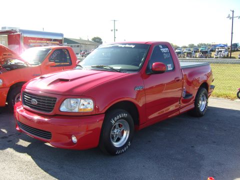 2007 Ford Mustang - A red Ford F-150 Lightning pickup truck, likely from the early 2000s, is parked next to another truck at a racing event.