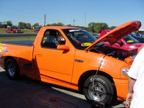 2007 Ford Mustang - A modified orange pickup truck is parked at a raceway with its hood open, showing off its engine and racing modifications.