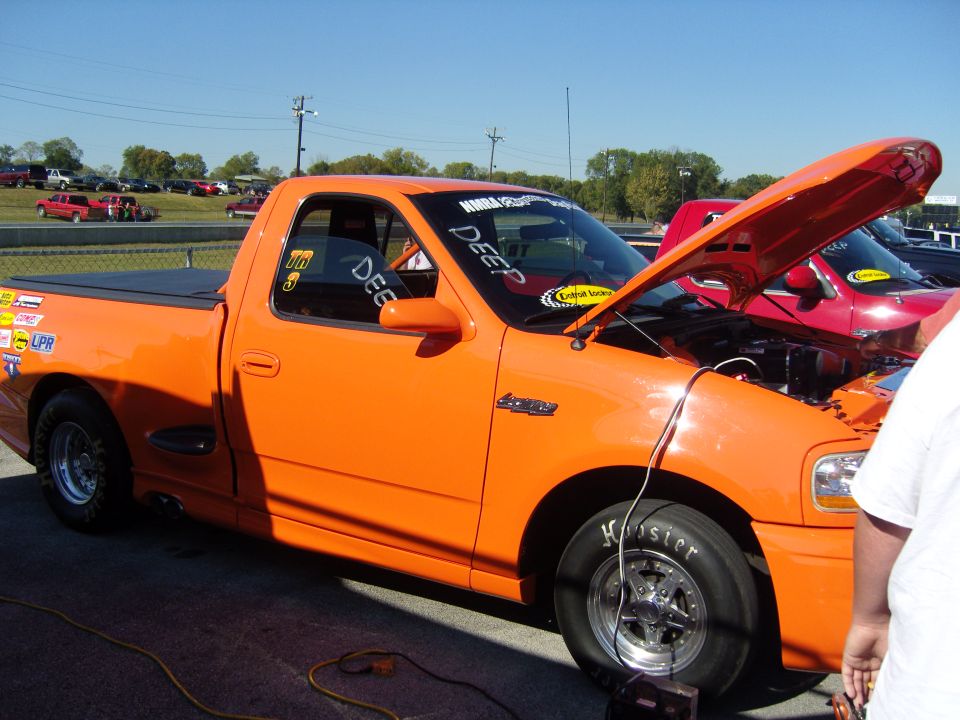 Orange pickup truck with hood open, racing tires, and various stickers. Another red vehicle is visible behind it