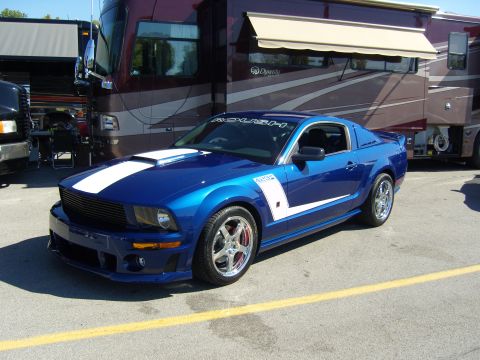 2007 Ford Mustang - A blue Ford Mustang with white racing stripes is parked in front of an RV.