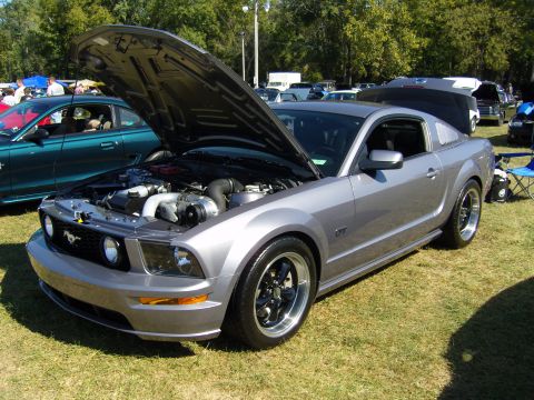 2007 Ford Mustang - A silver 2007 Ford Mustang GT is displayed with its hood open, revealing a supercharged engine at an outdoor car show.