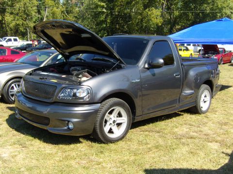 2007 Ford Mustang - A dark gray Ford F-150 SVT Lightning pickup truck is parked on a grassy field with its hood open, alongside other vehicles.