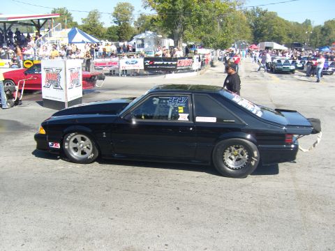 2007 Ford Mustang - A black drag car sits on the asphalt with a crowd watching in the background at a racing event.