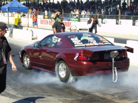2007 Ford Mustang - A maroon Ford Mustang drag car is seen performing a burnout on a race track, with spectators in the background.