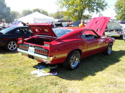 2007 Ford Mustang - Classic red car with gold side stripe and open trunk at an outdoor car show.