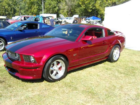 2007 Ford Mustang - A red 2007 Ford Mustang GT with black racing stripes is parked on a grassy field at an outdoor event.