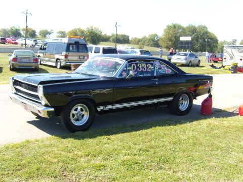 2007 Ford Mustang - A black 1960s Ford Fairlane is parked on the grass at a race track, with a red gas can nearby.