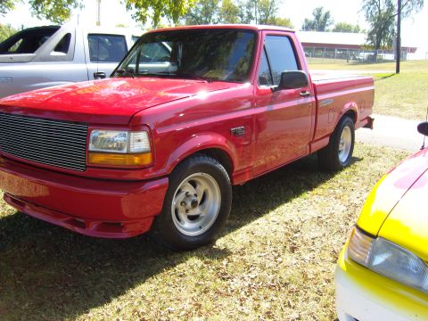 2007 Ford Mustang - Red pickup truck with aftermarket wheels and chrome grille. Another vehicle is visible behind it.