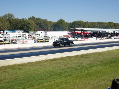 2007 Ford Mustang - A black pickup truck races down a drag strip with grandstands full of spectators.