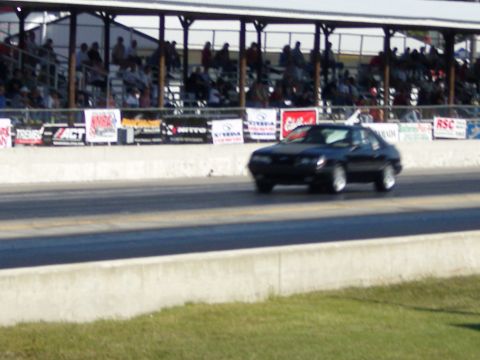 2007 Ford Mustang - A black coupe speeds down a racetrack with spectators watching from the stands.