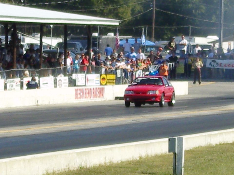 Red Fox body Mustang drag car on a race track, front view. Spectators in background
