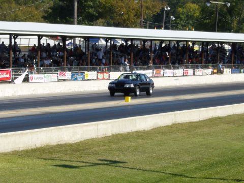 2007 Ford Mustang - A black coupe, likely a 1990s Ford Mustang, races down a drag strip with spectators watching.