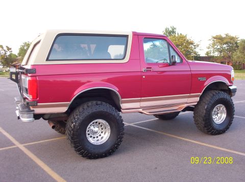 1995 Ford Bronco - A red 1995 Ford Bronco with a tan top is parked on asphalt. The vehicle has large off-road tires and chrome wheels, and a spare tire rack i…