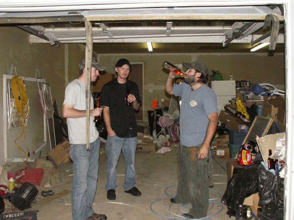 Three men standing in a cluttered garage, two holding beer bottles, one drinking from a bottle.