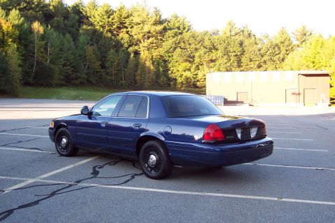 2001 Ford Crown Victoria - A blue sedan, possibly a former police vehicle, is parked in an empty lot with trees and a building in the background.