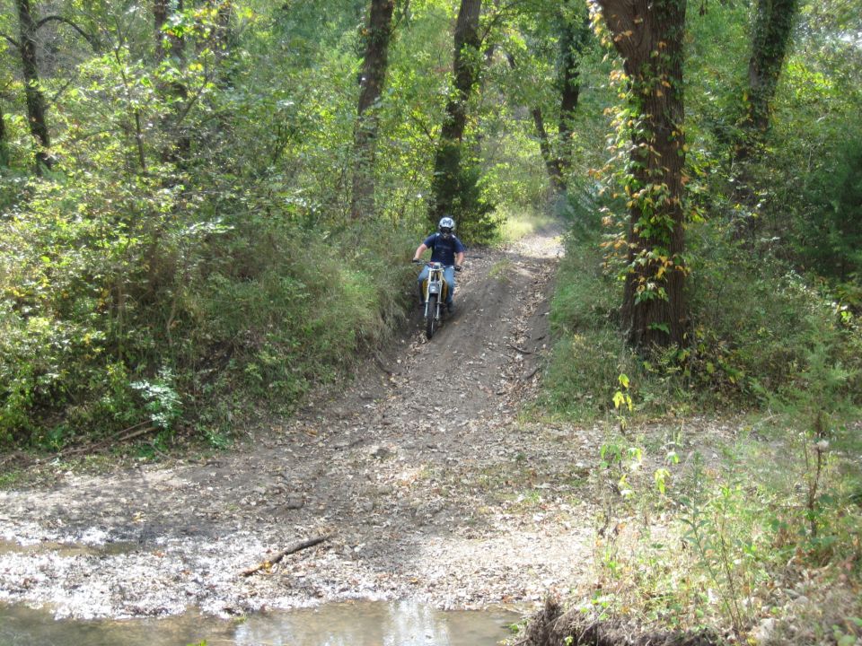 Dirt bike rider on a gravel path through trees, with a small stream in the foreground