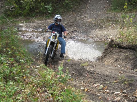 1993 Suzuki RMX250 - A rider navigates a water obstacle on a dirt bike, kicking up spray on a wooded trail.