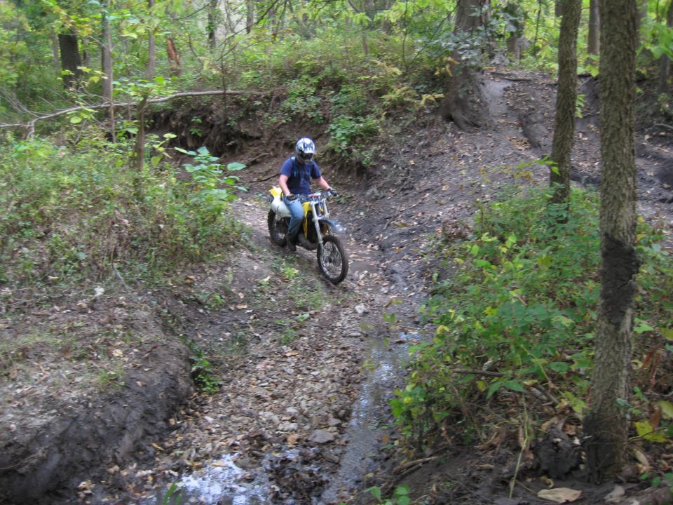Dirt bike with yellow plastic and a rider in a blue shirt and white helmet navigates a muddy, wooded trail