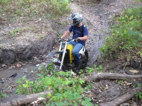 1993 Suzuki RMX250 - A rider on a dirt bike maneuvers through a challenging muddy terrain, surrounded by overgrown vegetation.