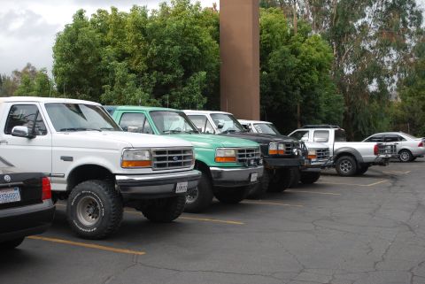 1996 Ford Bronco - A line of trucks and SUVs, including a white 1996 Ford Bronco, parked in a lot.