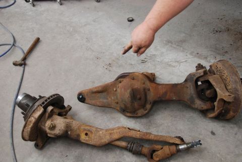 1996 Ford Bronco - Two heavily rusted front axle assemblies are laid out on a concrete floor, with a person's hand pointing towards them.