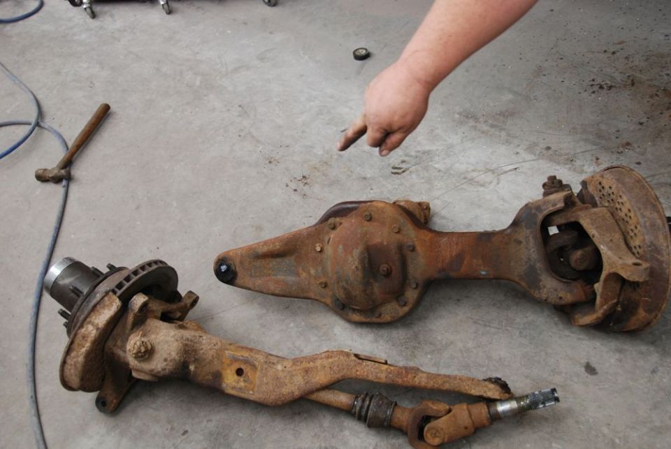 Two rusty front axle assemblies on a concrete floor, with a hand pointing at one
