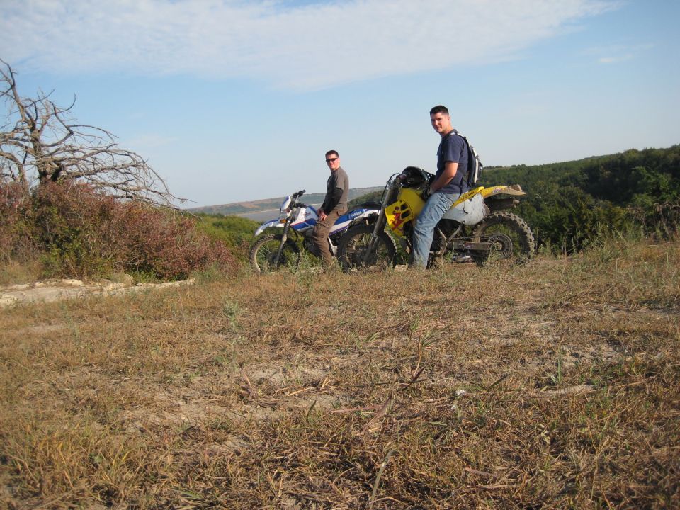 Two dirt bikes parked on a hill with riders. Dry grass and bushes surround them